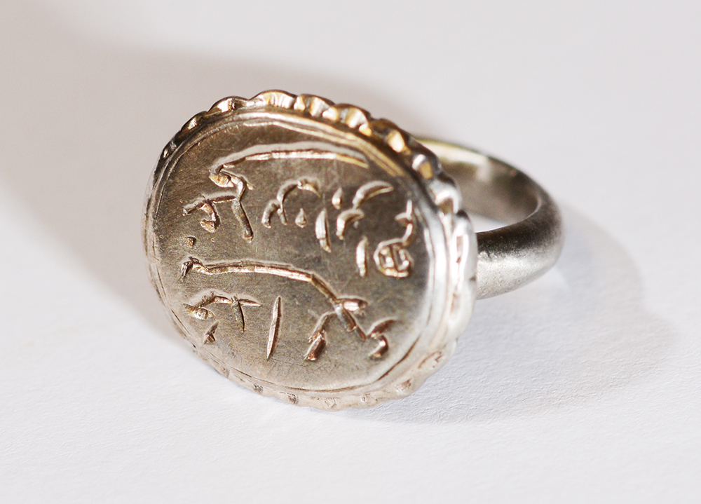 Öntött ezüst gyűrű, palástjának szélén körben hullámvonalas díszítés található, ovális alakú pecsétlő részén vésett felirattal